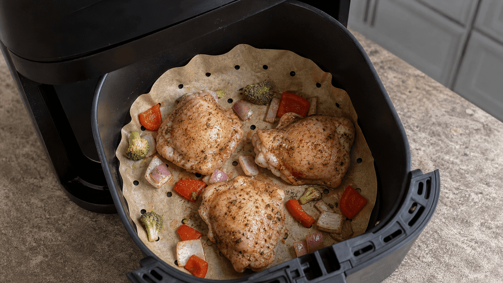 Perforated parchment held down by chicken and vegetables inside an air fryer basket