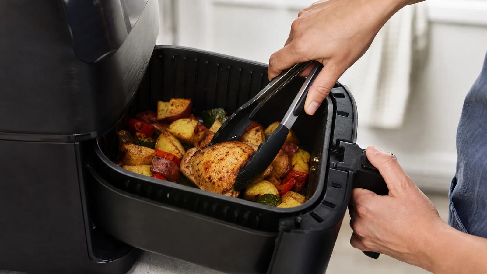 A person turning chicken and vegetables with tongs in an open air fryer basket